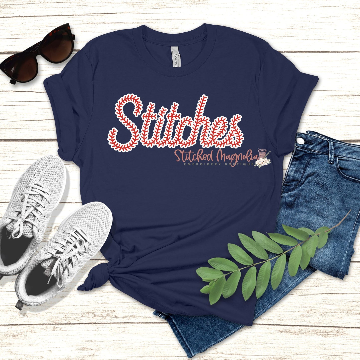 Navy t-shirt with the word Stitches in red baseball-stitch script lettering with a white outline, styled flat lay with sneakers and jeans