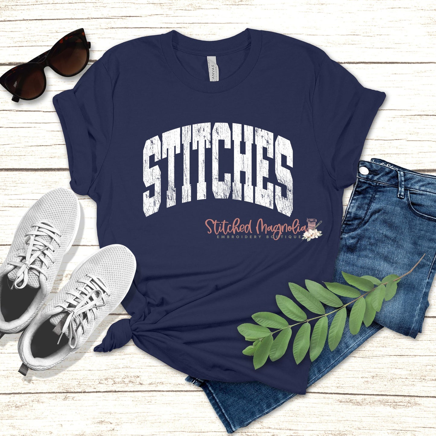 Navy t-shirt with distressed white varsity lettering spelling STITCHES arched across the chest, styled flat lay with sneakers and jeans