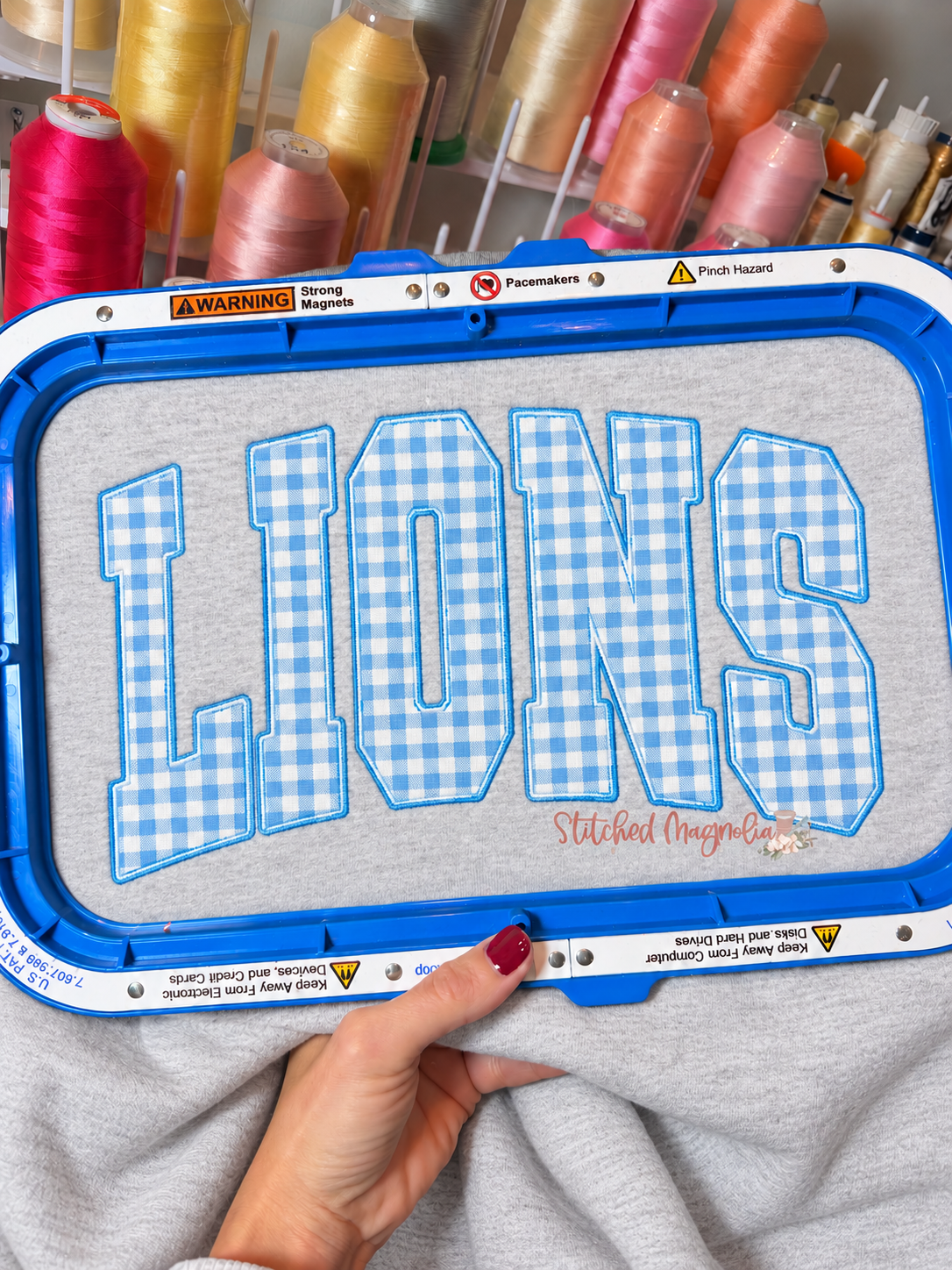 Ash grey sweatshirt in an embroidery hoop with blue-and-white gingham LIONS fabric appliqué and blue embroidered outlines.